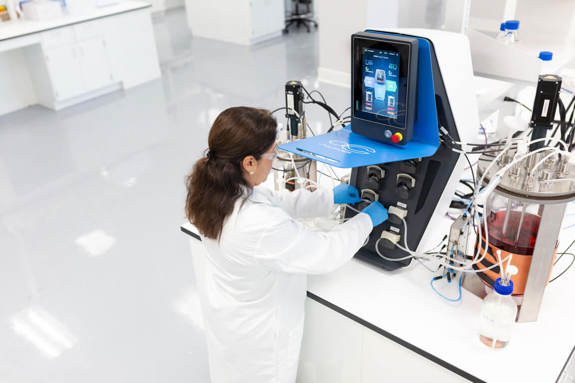 Scientist operating AUXO V benchtop bioreactor and adjusting tubing connections in laboratory.