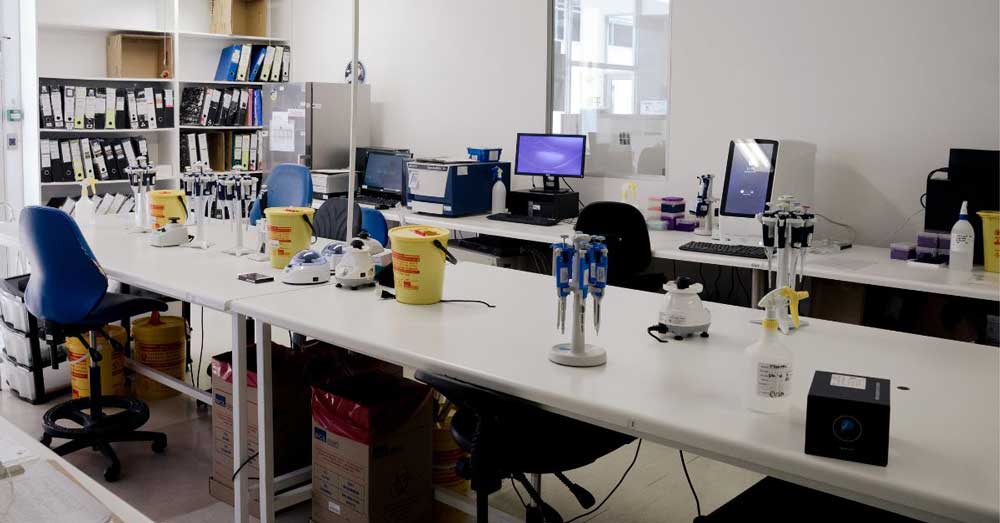 Clean laboratory workspace with white benches, pipettes, biohazard bins, and computers ready for lab work.